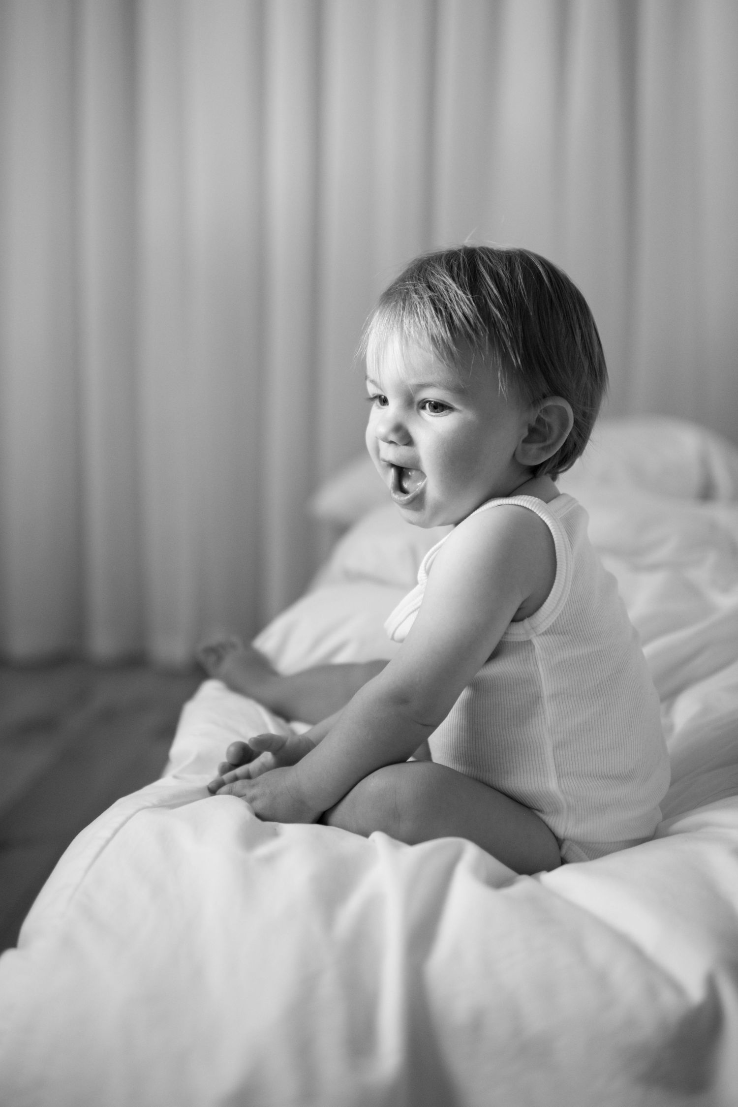 Black and white photo of a happy toddler sitting on a white bed, laughing and wearing a sleeveless white outfit.