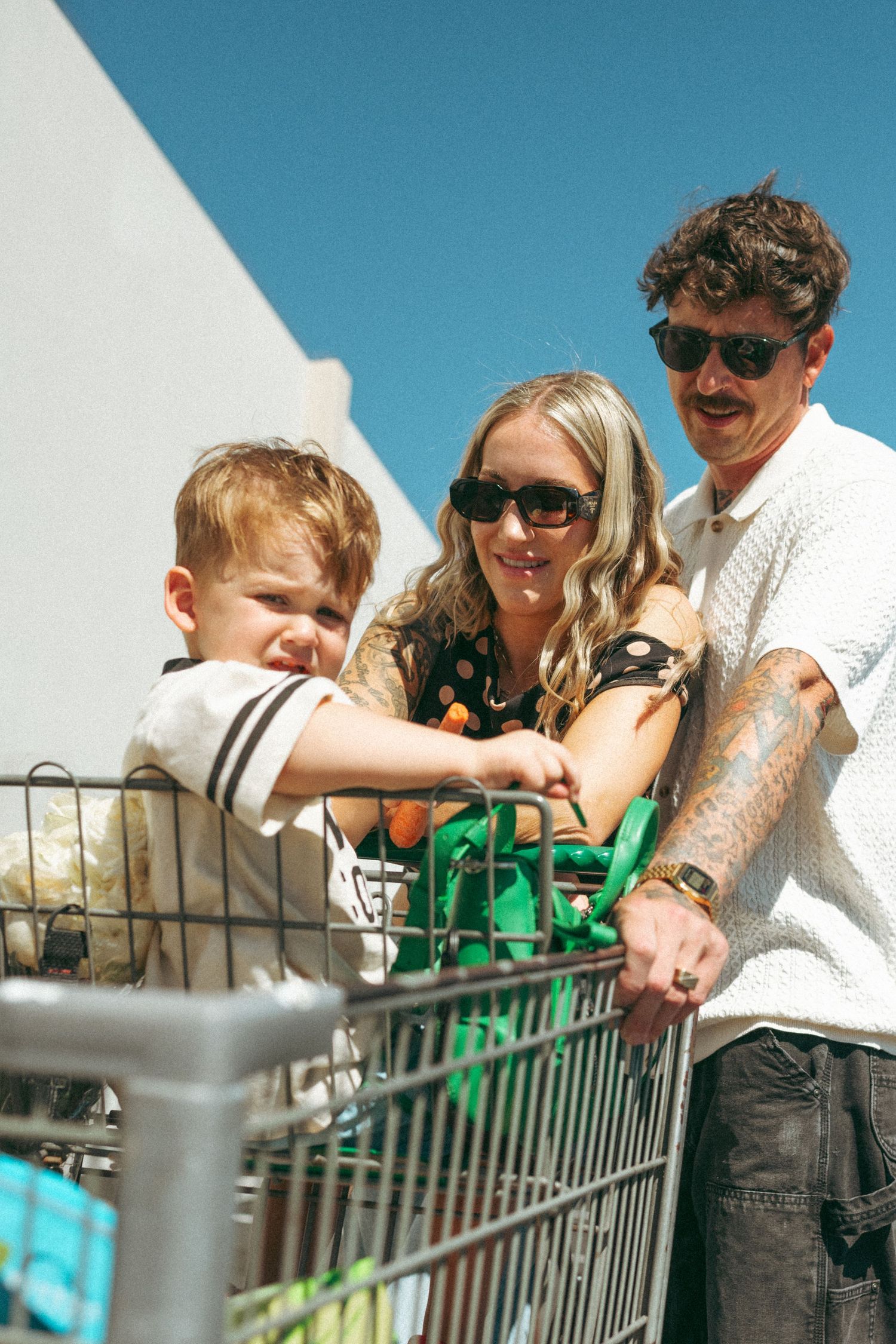 Young family with toddler in shopping cart outside on sunny day, parents wearing sunglasses.