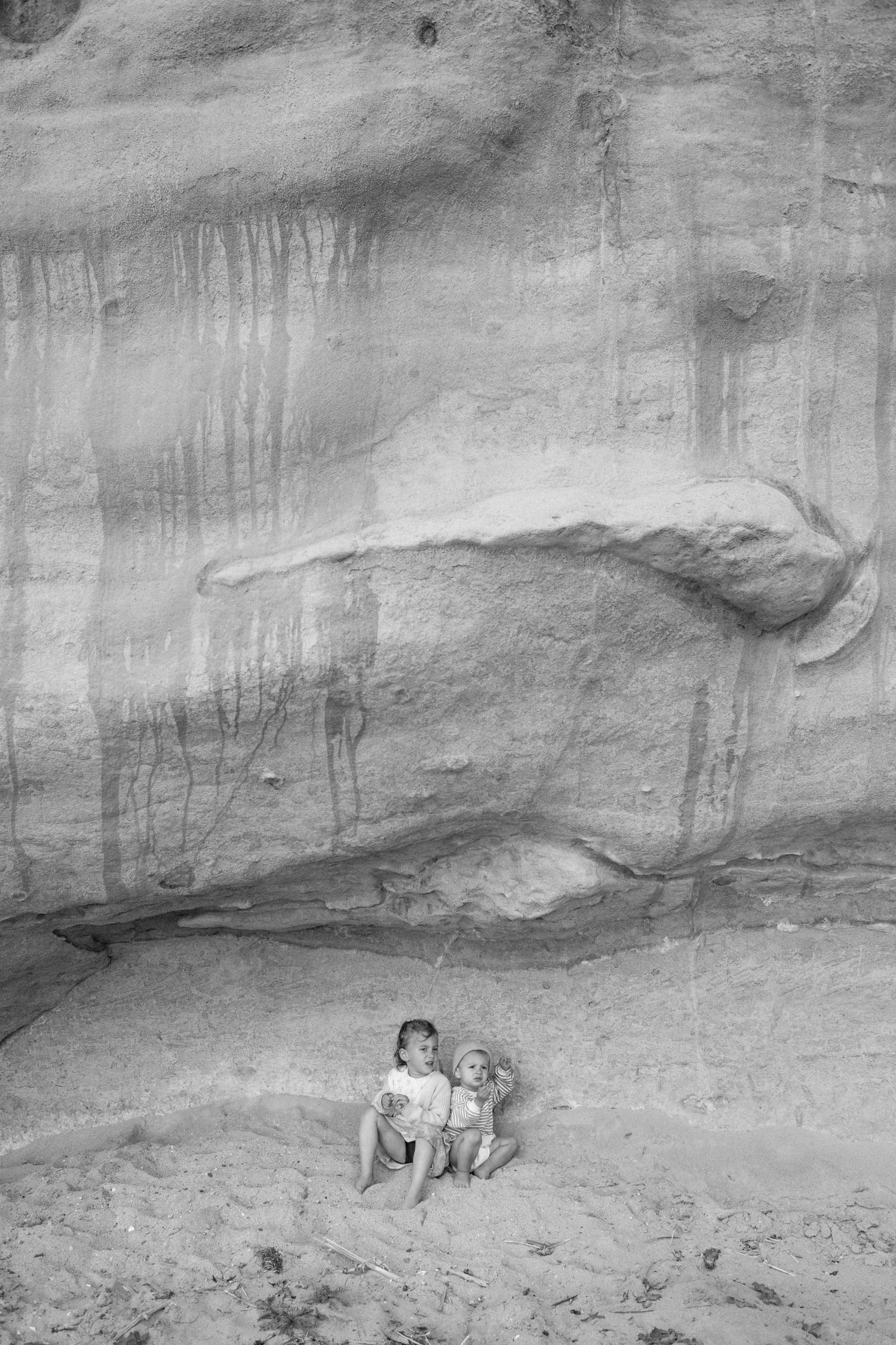 Two young children sit together in the sand beneath a dramatic sandstone rock formation in black and white.