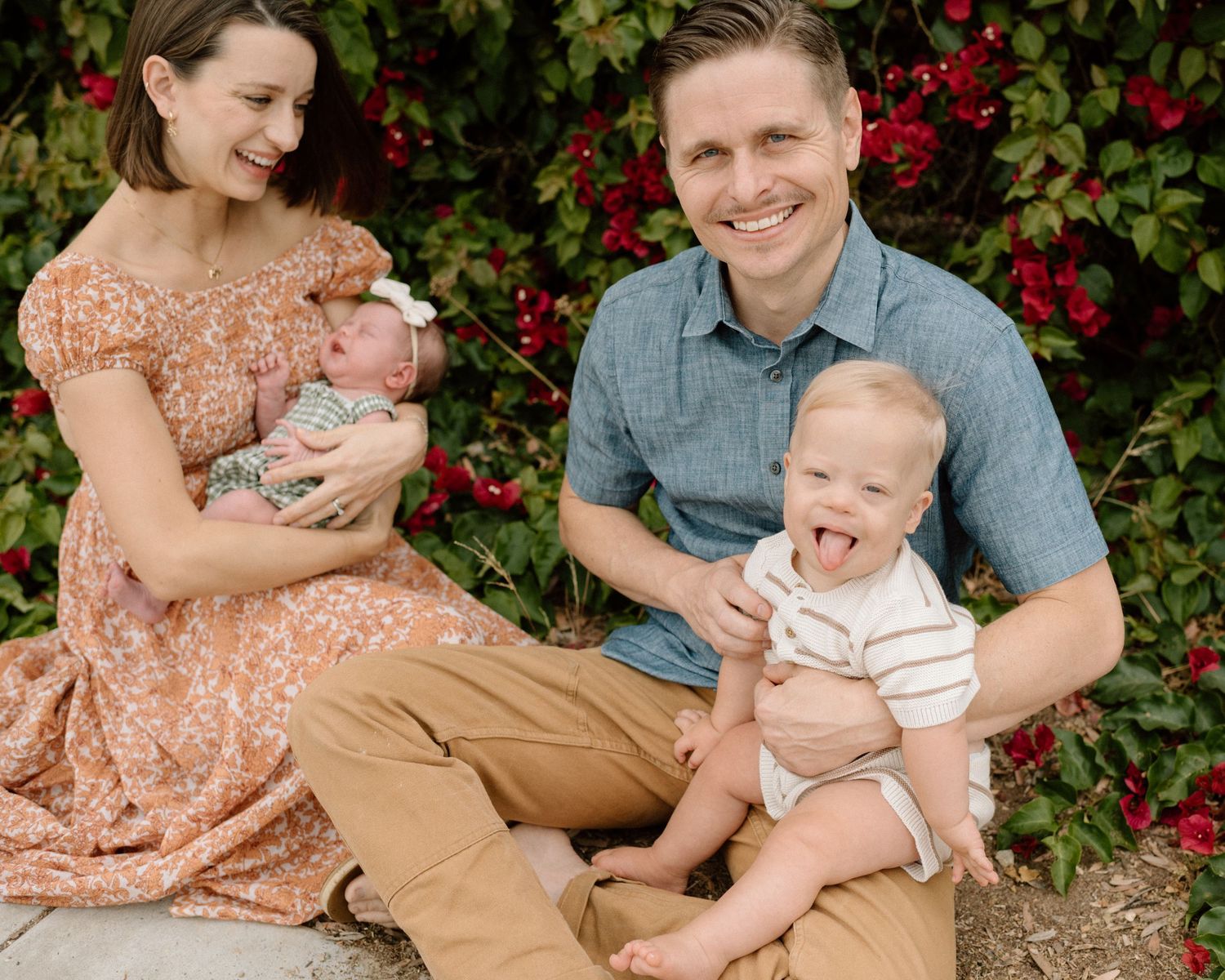 Smiling family of four with newborn and toddler posing outdoors near red flowers.