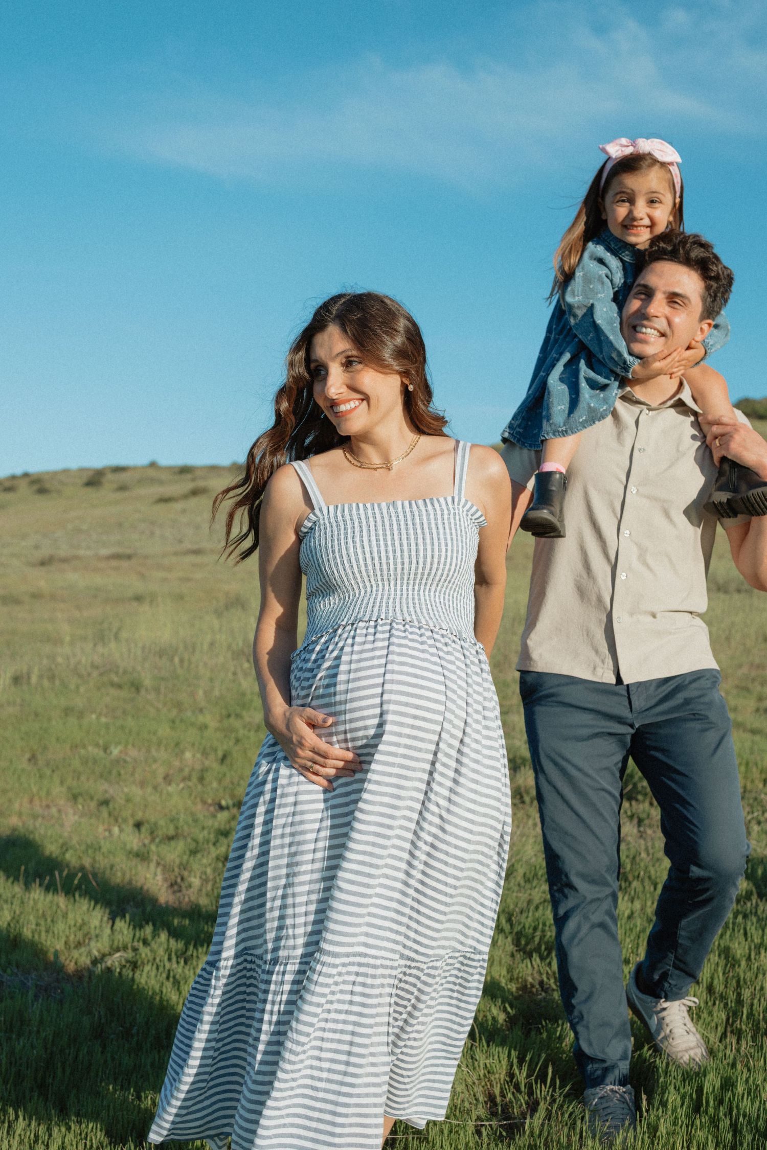 Happy pregnant family with daughter on fathers shoulders in green field.