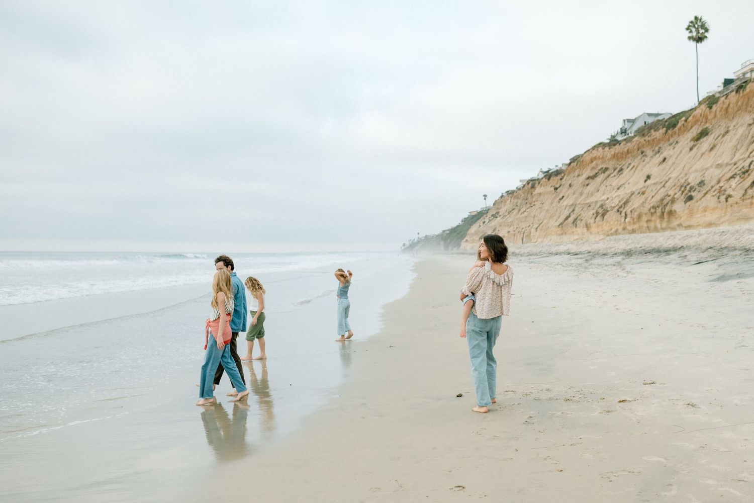 A family of six walking and playing along a sandy beach with dramatic cliffs and ocean waves in the background.