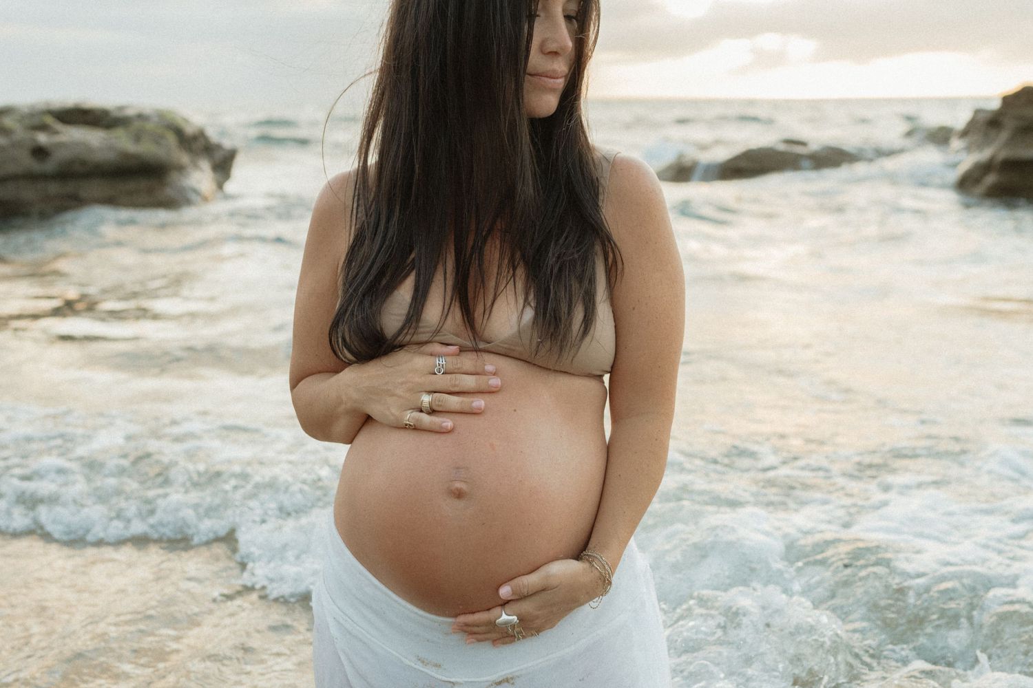 Pregnant woman in white wrap holding baby bump on rocky beach with ocean waves at sunset.