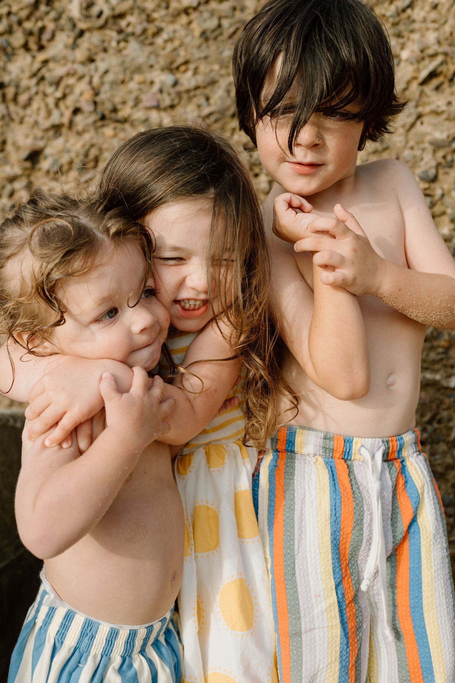 Three happy children hugging and laughing together outdoors in summer clothing.