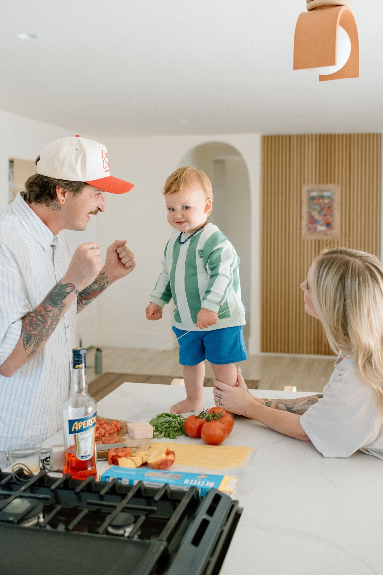 Family cooking together in modern kitchen with toddler standing on counter next to stove and fresh ingredients.