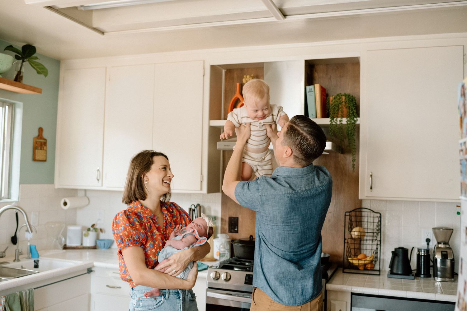 Young family laughing in white kitchen, father lifting baby while mother holds newborn.