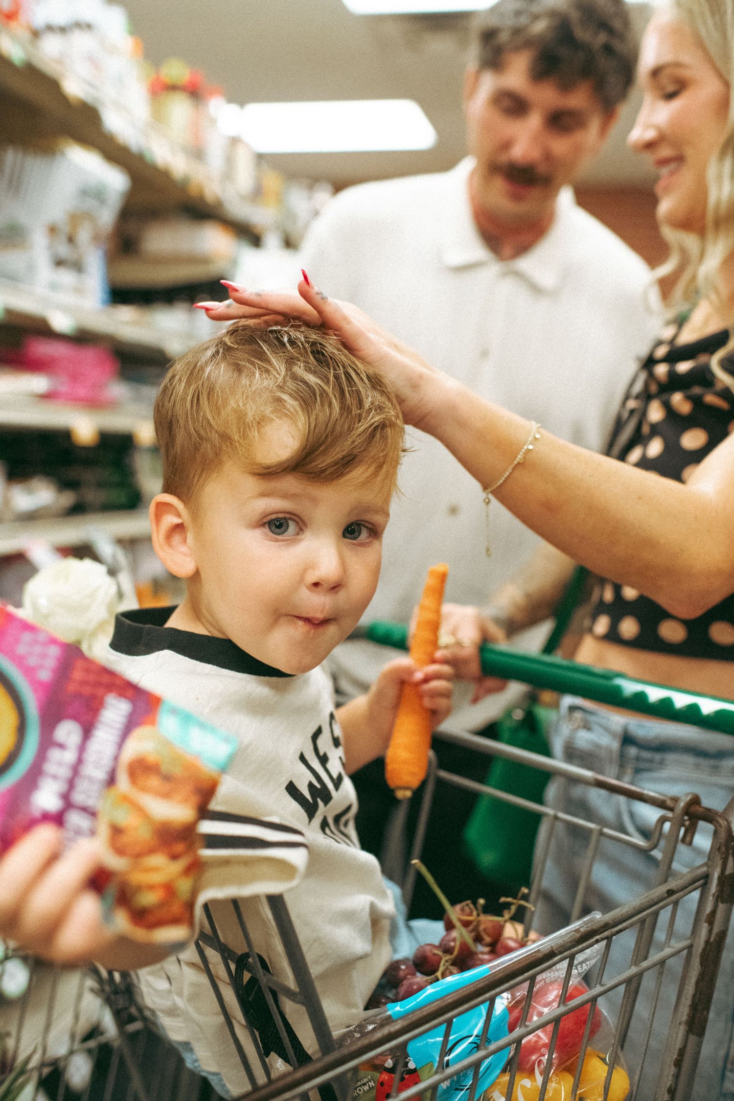 Young boy sitting in shopping cart holds snack while mother strokes his hair in grocery store aisle.