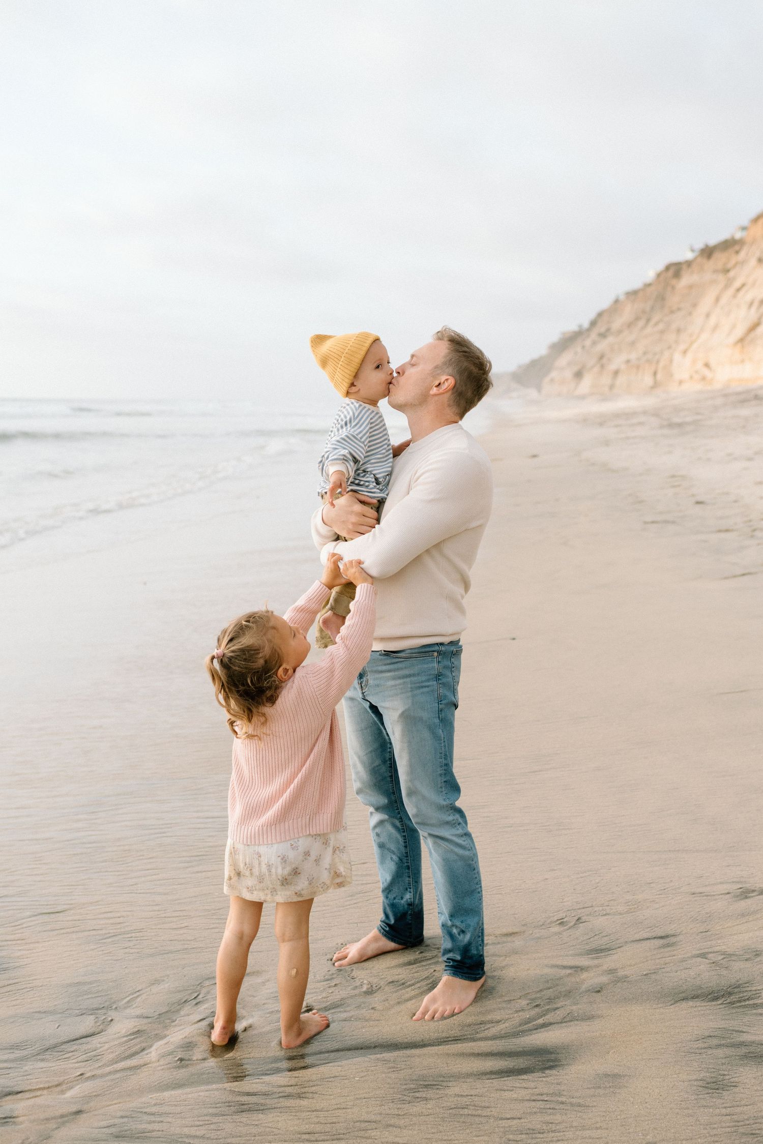 Father kissing baby while daughter holds his hand on a sandy beach at sunset.