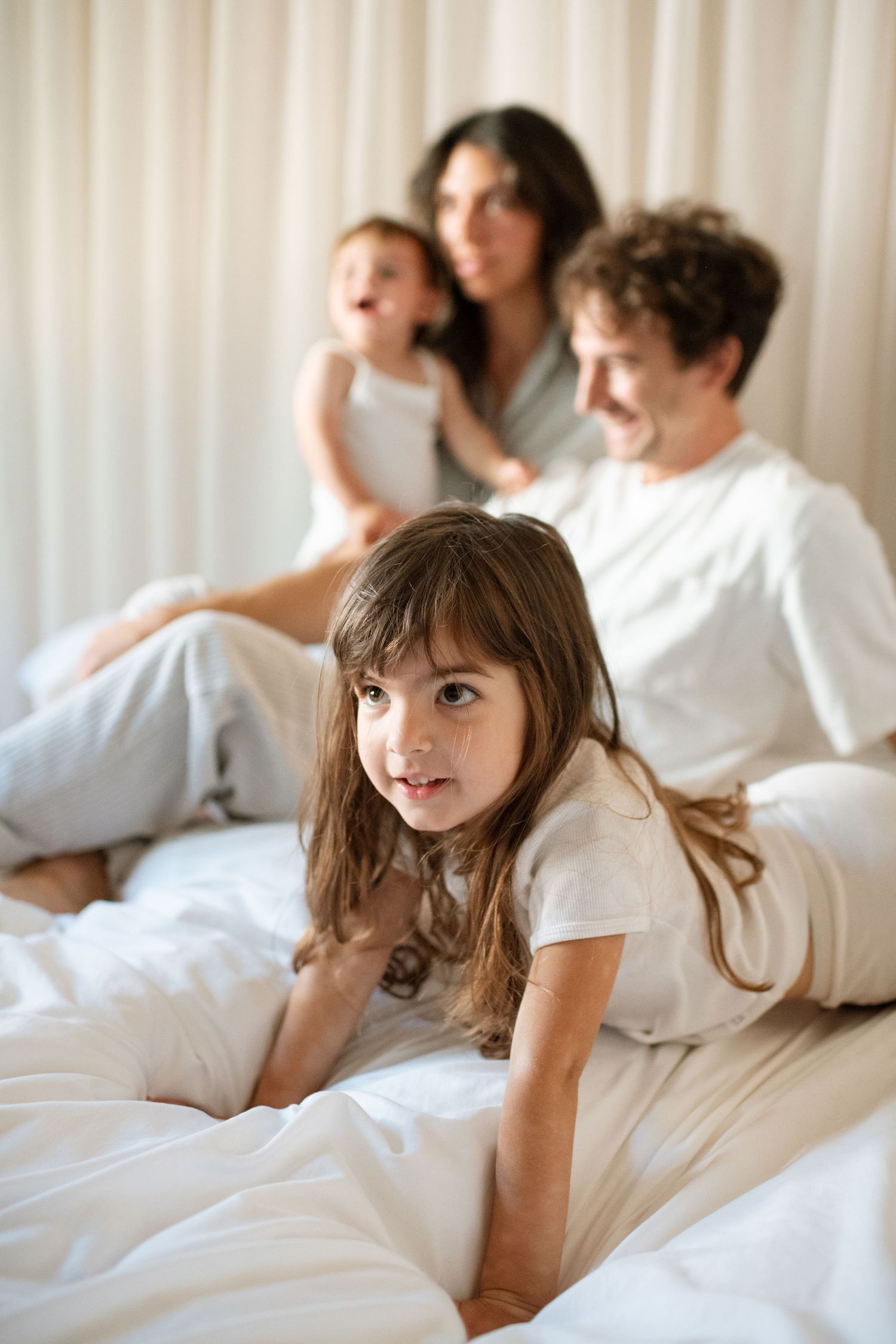 A young girl lies on a white bed in the foreground while her family sits and smiles together in the background.