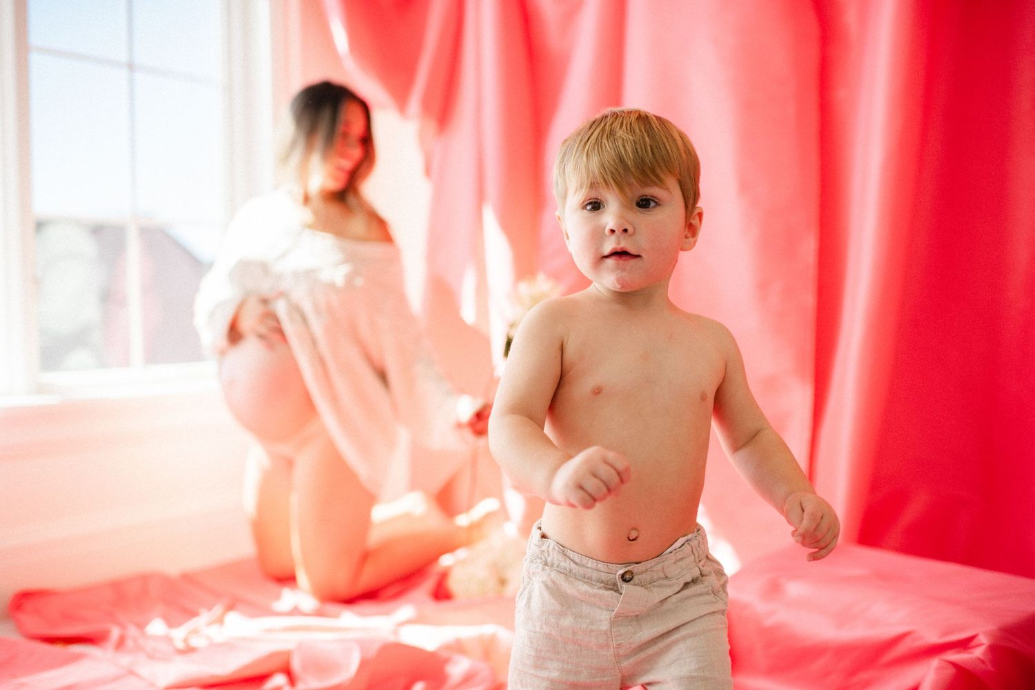 Toddler boy standing shirtless in front of red curtains with pregnant woman posing in soft natural light background.