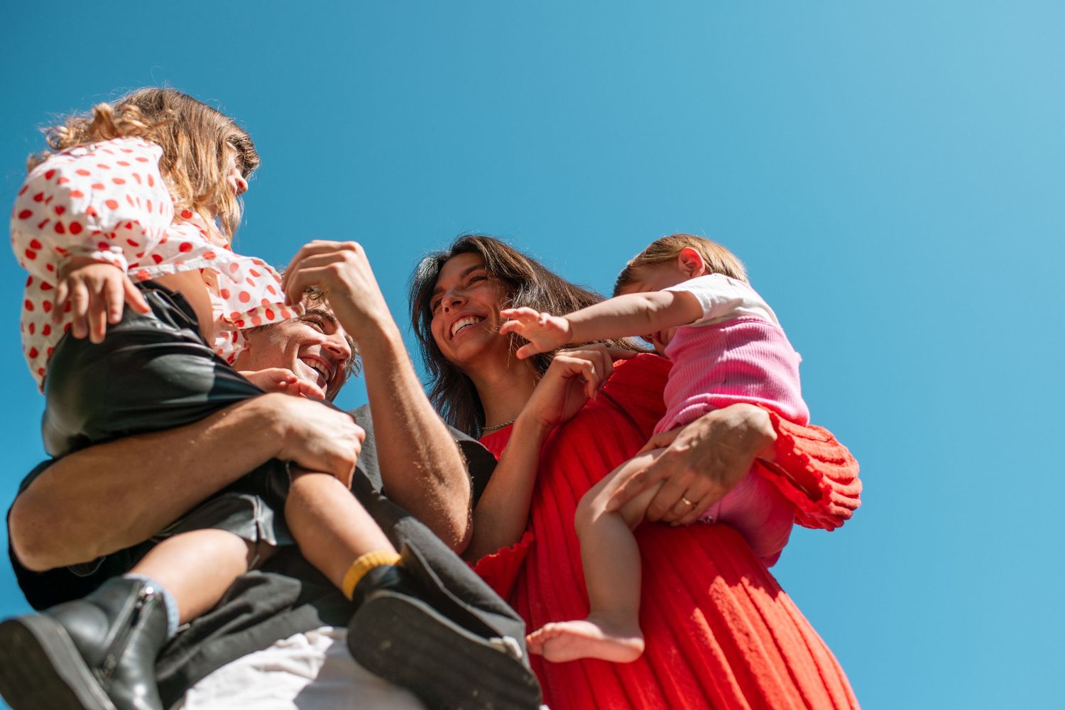 Family with two young children smiling and making heart shapes against a bright blue sky on a sunny day.