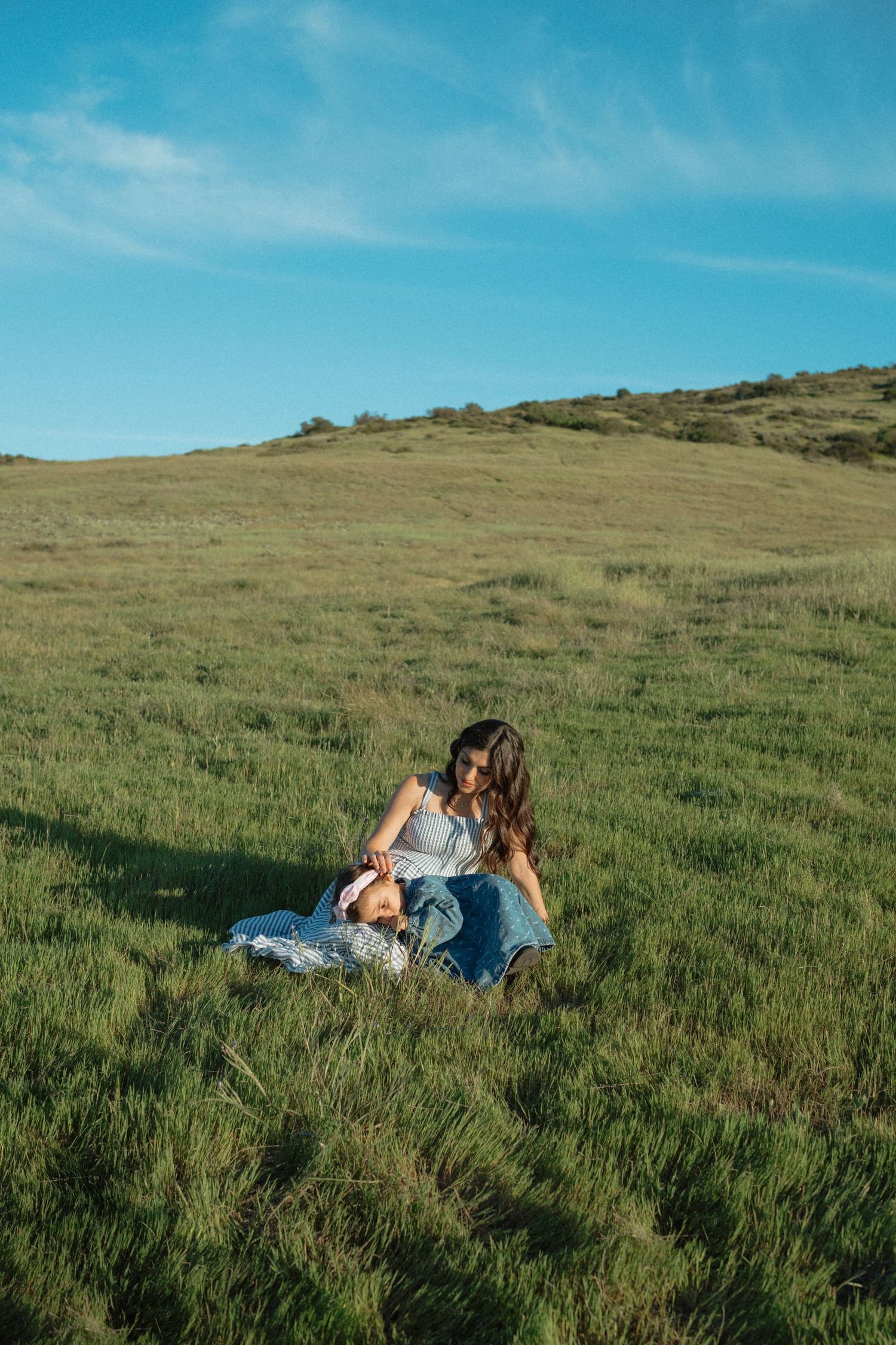 Woman in white top and jeans sitting with baby in lush green field under blue sky.