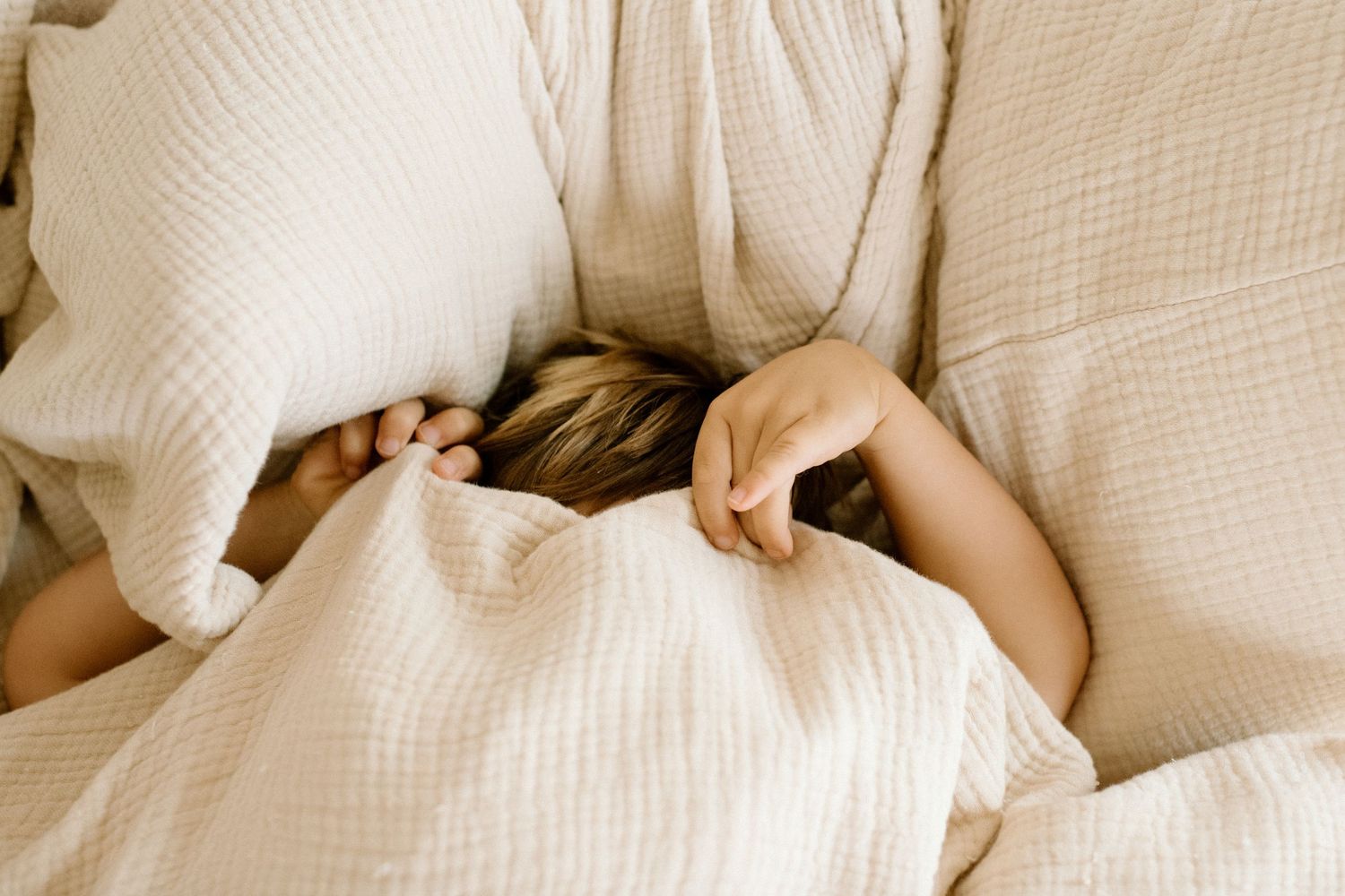 Person hiding under cozy white waffle-knit bedding and pillow in a soft, neutral bedroom setting.