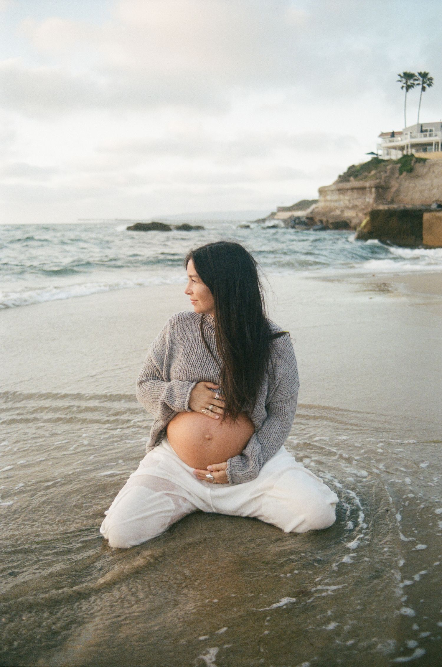 Pregnant woman in gray sweater and white pants sitting on beach shore cradling her baby bump.