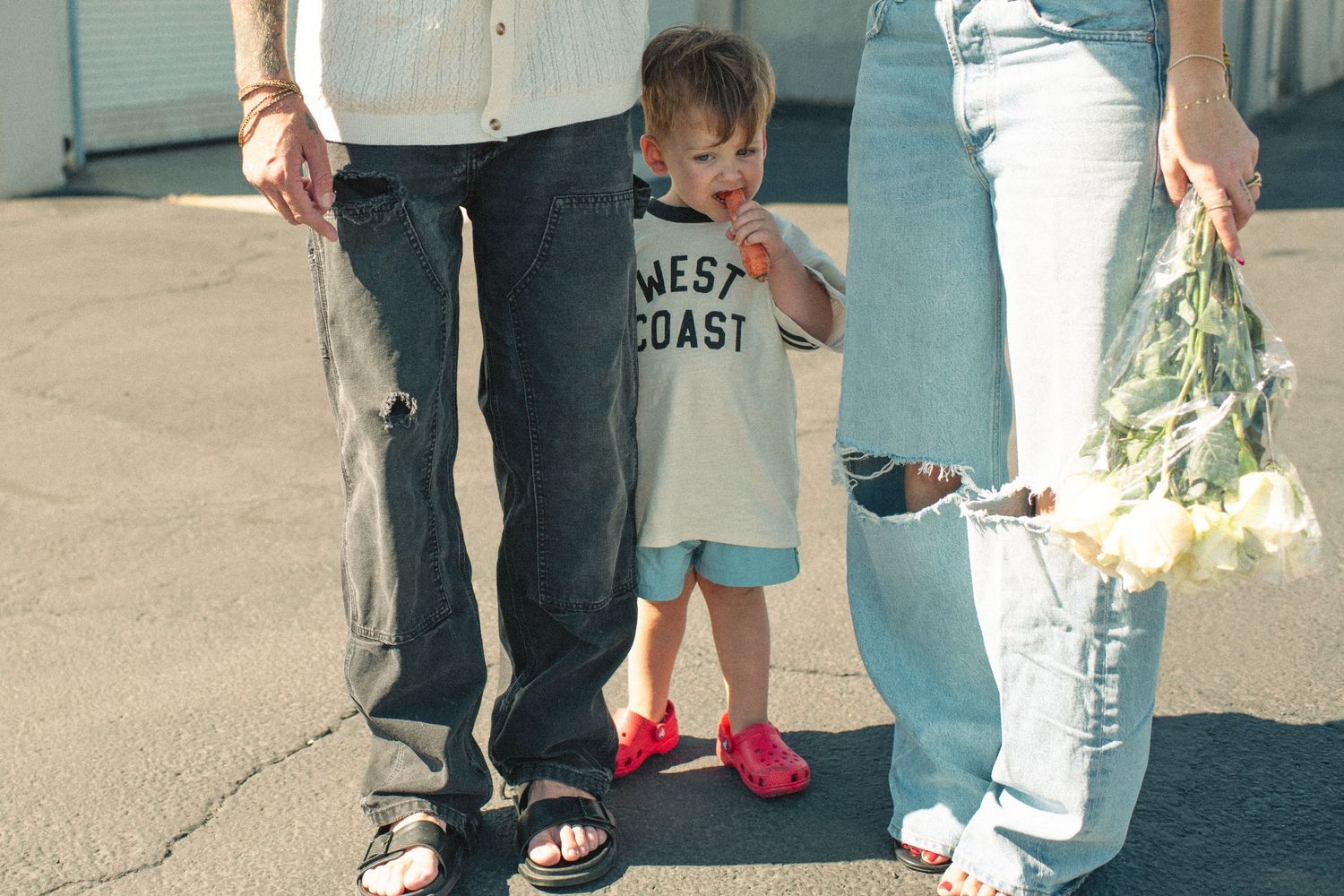 Toddler in West Coast shirt and pink shoes stands between two adults in jeans, one holding white flowers.