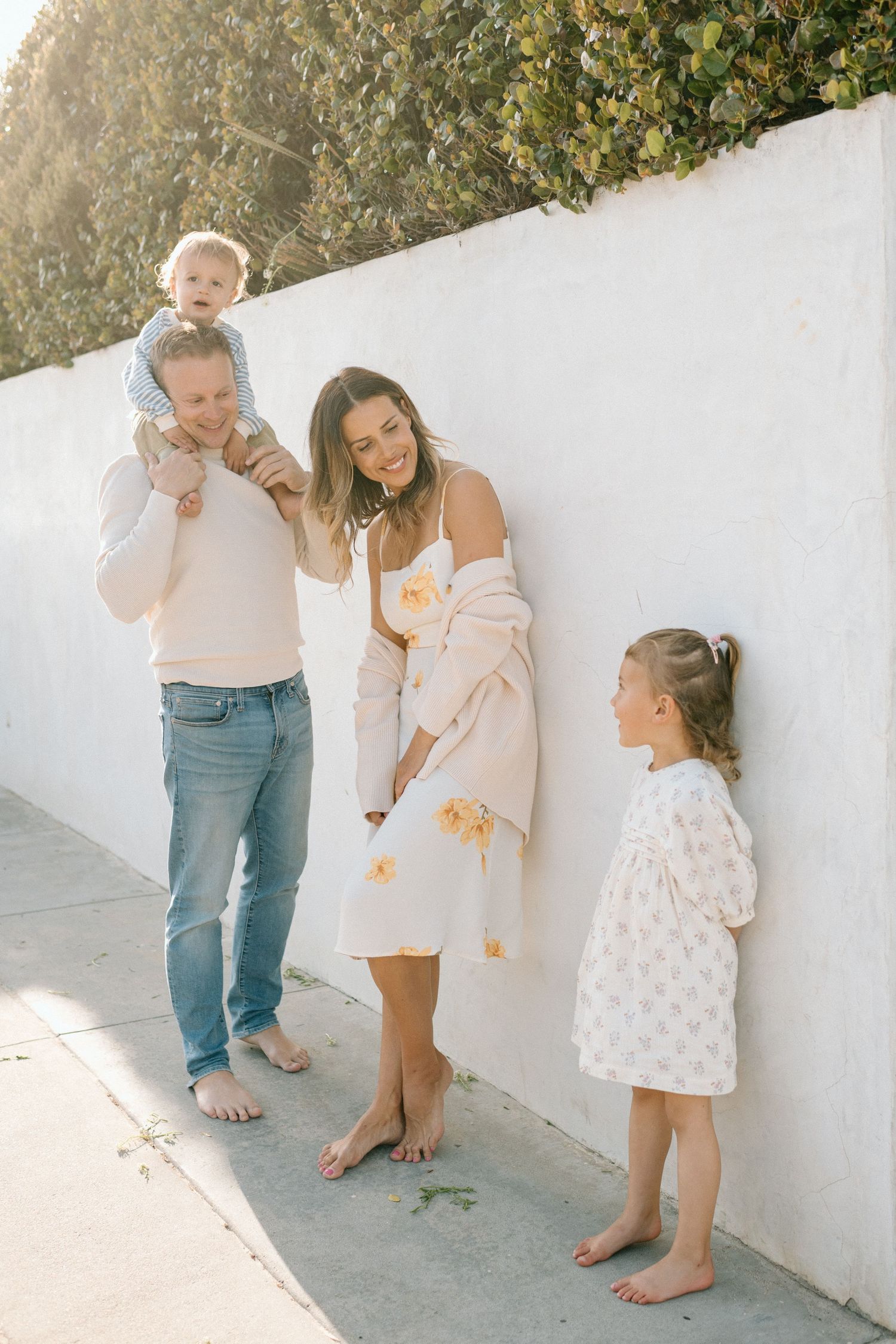 Family of four in neutral tones standing barefoot against white wall in golden sunlight.