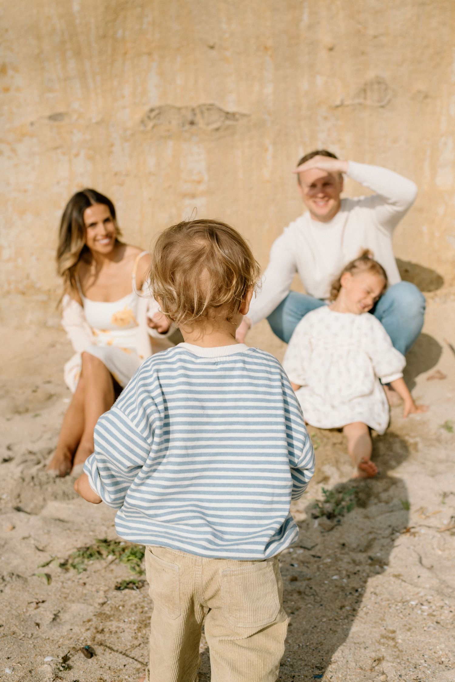Family relaxing on sandy beach, two adults sitting against wall with toddler while young child walks toward them.