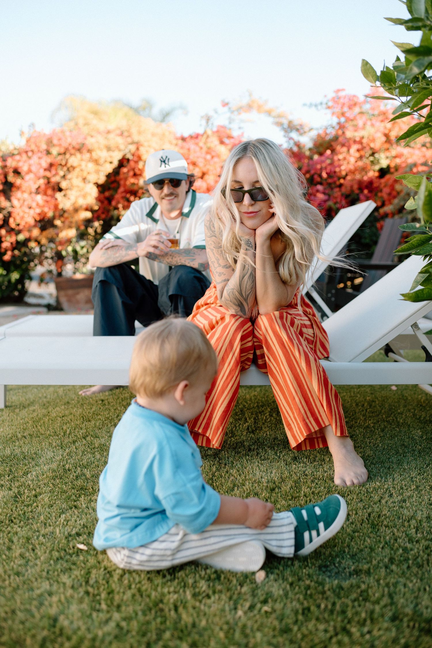 Stylish couple with baby relaxing on lawn chairs surrounded by vibrant bougainvillea blooms.