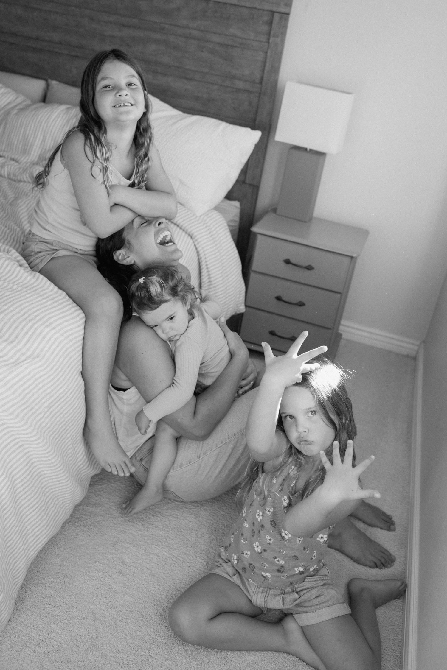 Mother and three children playfully piling together on bedroom floor in black and white photo.
