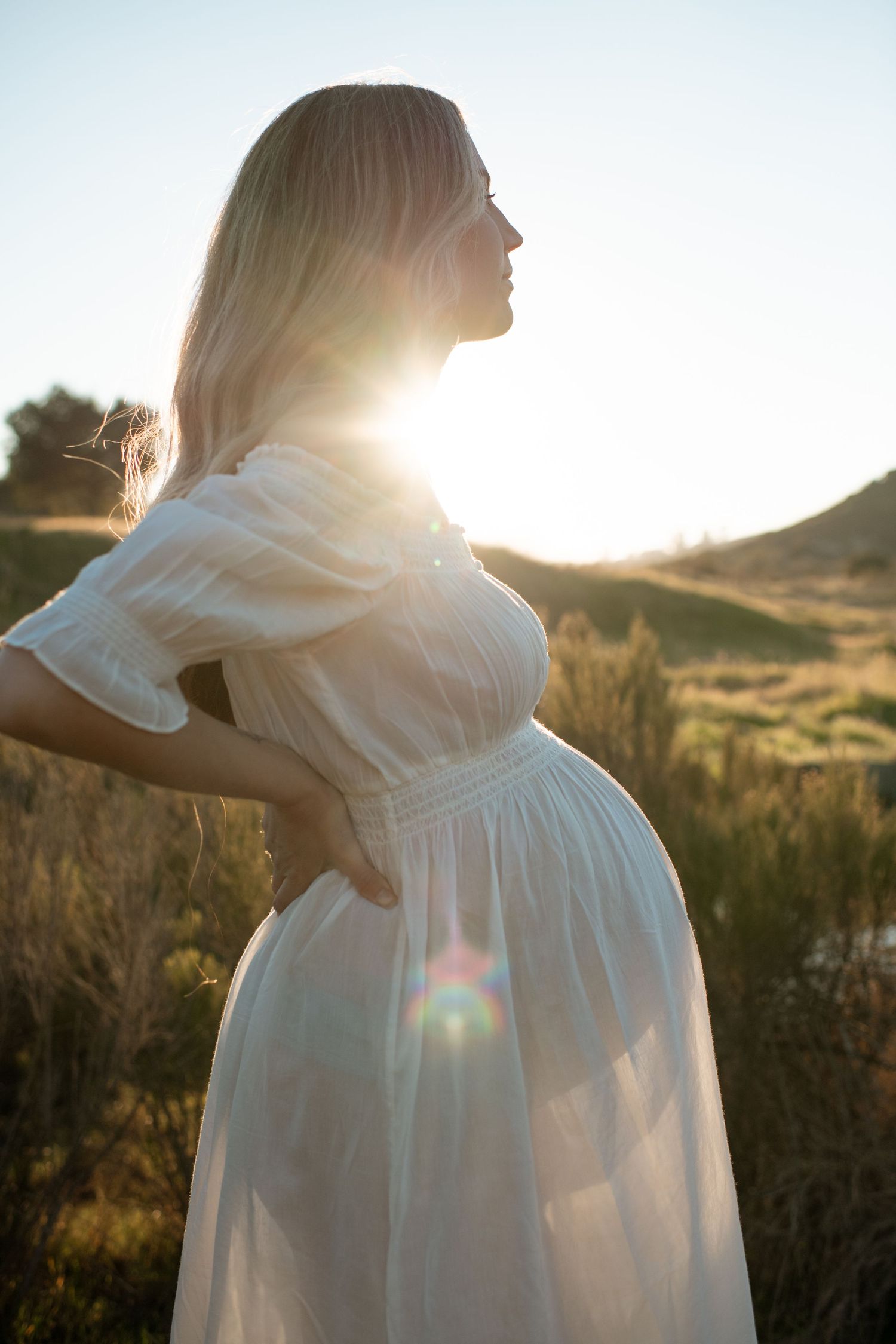 Pregnant woman in white dress standing in golden sunlight at sunset in a natural field setting.