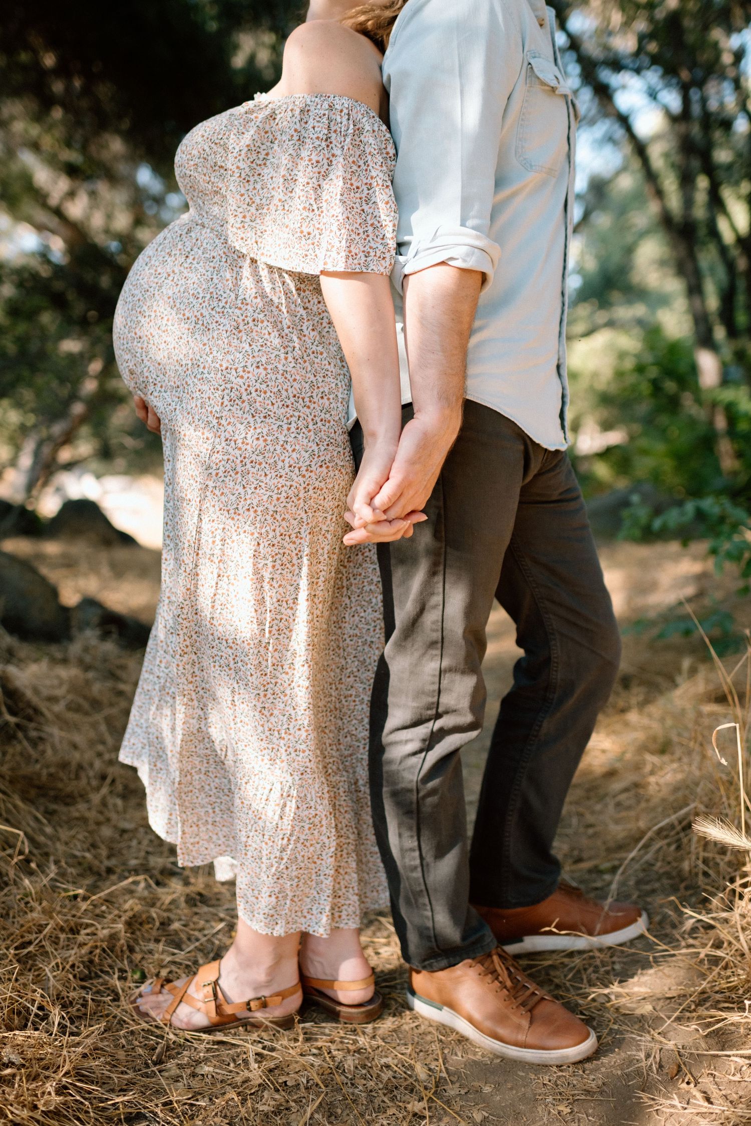 Pregnant woman in floral off-shoulder dress holding hands with partner outdoors.