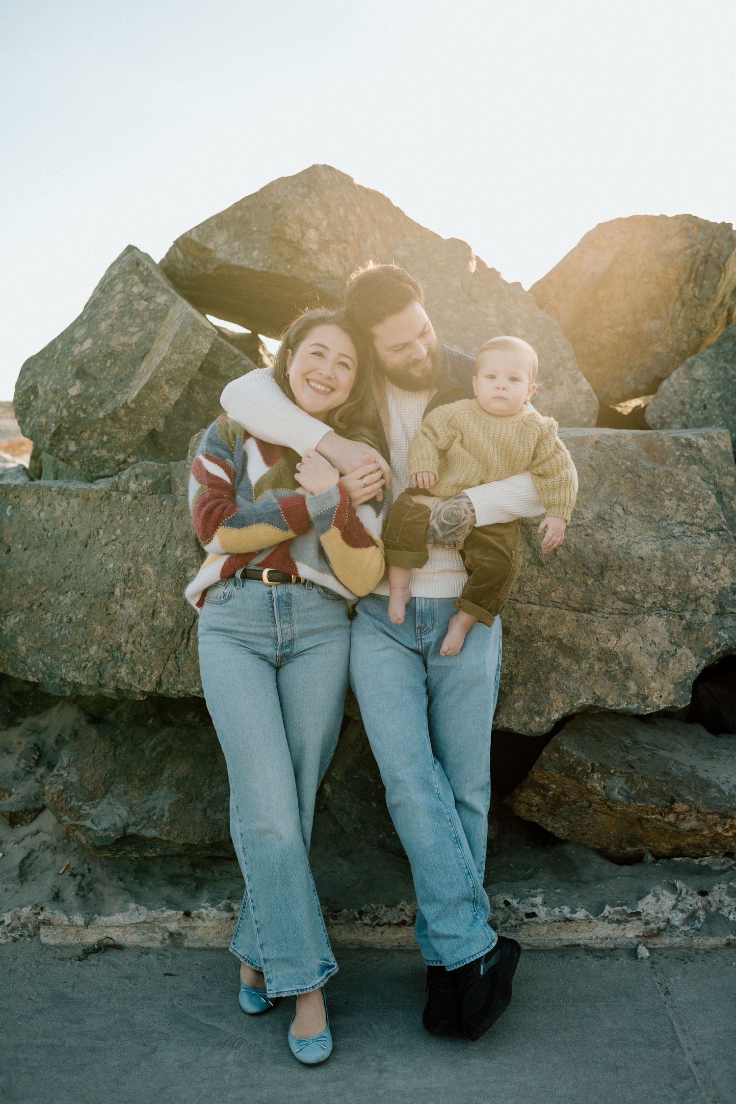 A happy young family with a baby in a yellow sweater poses against large rocks in warm sunlight.