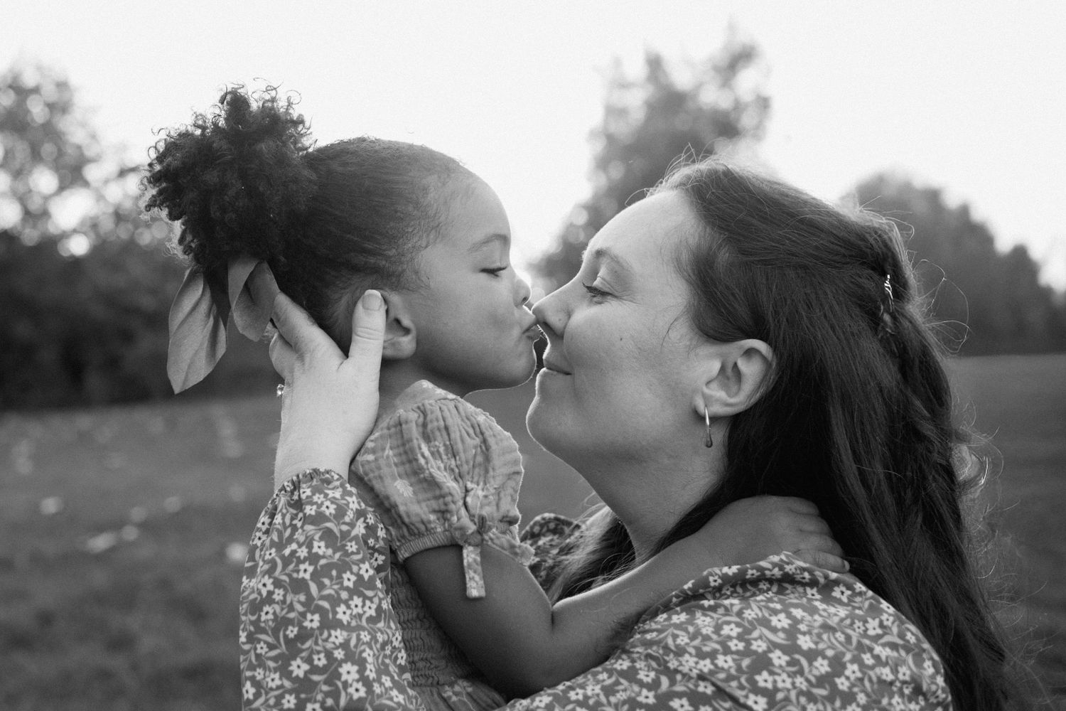 Black and white photo of young girl giving her mother a tender kiss on the nose outdoors.