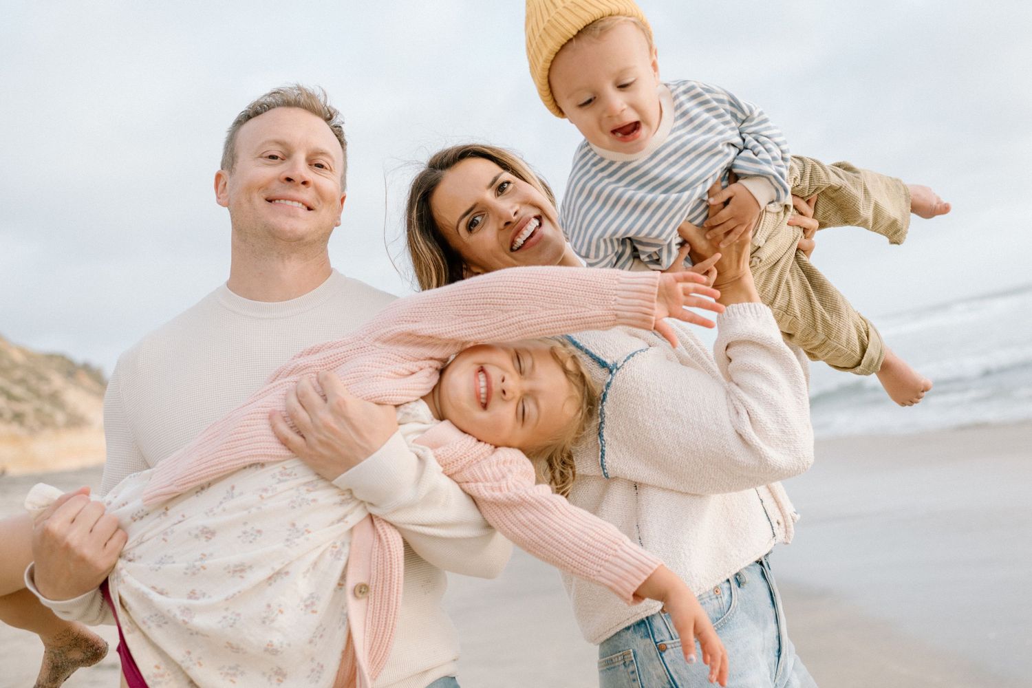 Happy family of four enjoying a beach day, parents playfully holding young children with ocean backdrop.