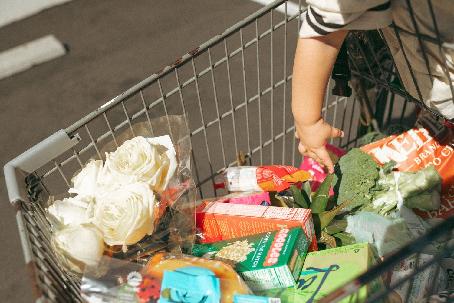 Overhead view of a full grocery cart with fresh flowers, vegetables, and packaged foods being shopped outdoors.