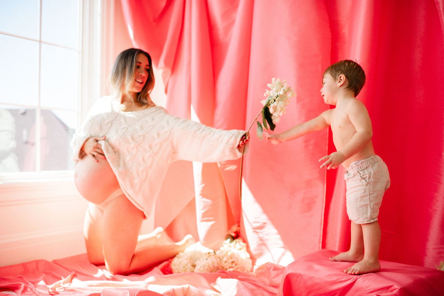 Pregnant woman in white sweater kneeling as young boy offers her flowers on pink backdrop.
