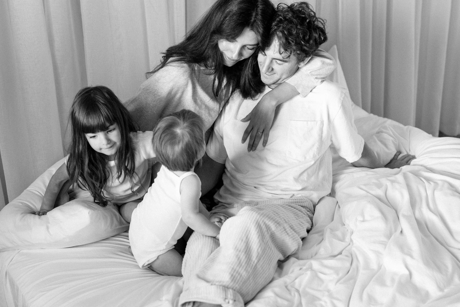 Black and white family photo of parents and two young children sitting together on a bed in cozy white clothing.
