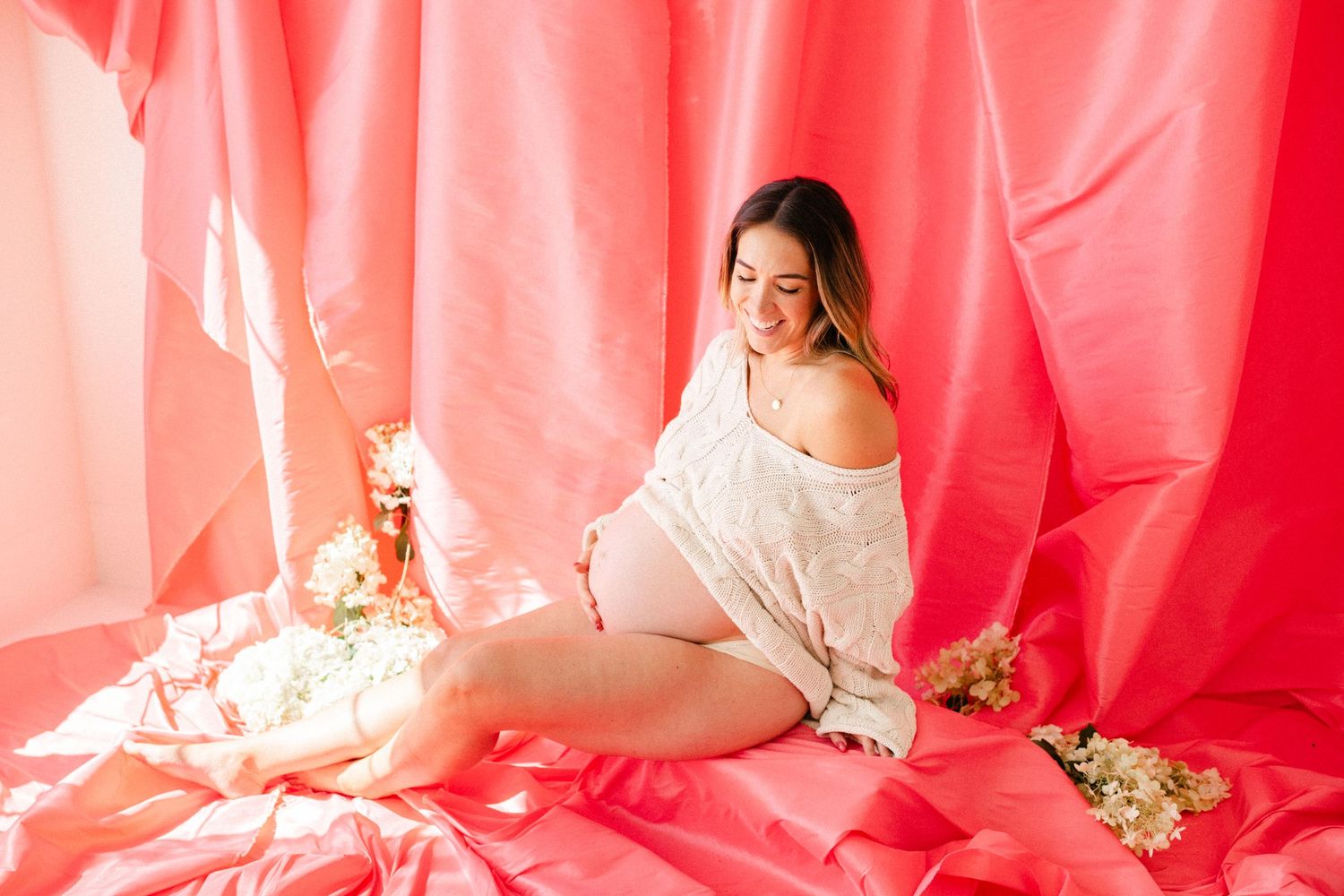 Smiling pregnant woman in white lace top sits on pink fabric backdrop surrounded by delicate white flowers in bright studio.