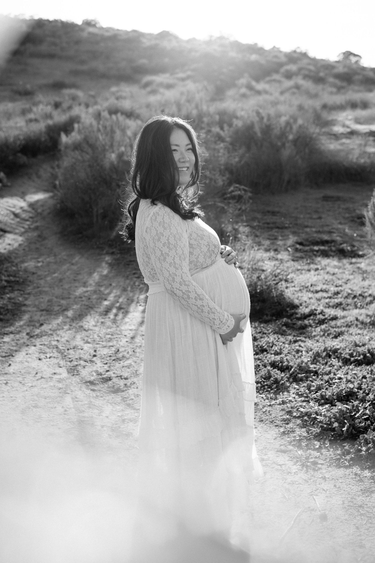 Black and white maternity portrait of smiling pregnant woman in white lace dress at golden hour.