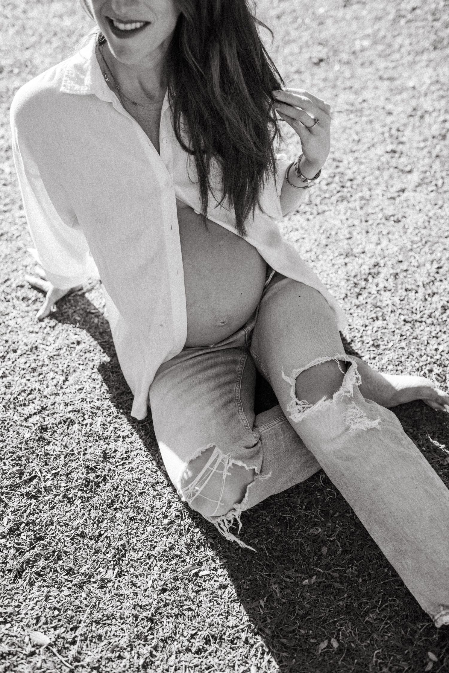 Black and white photo of pregnant woman in white shirt and ripped jeans sitting outdoors.