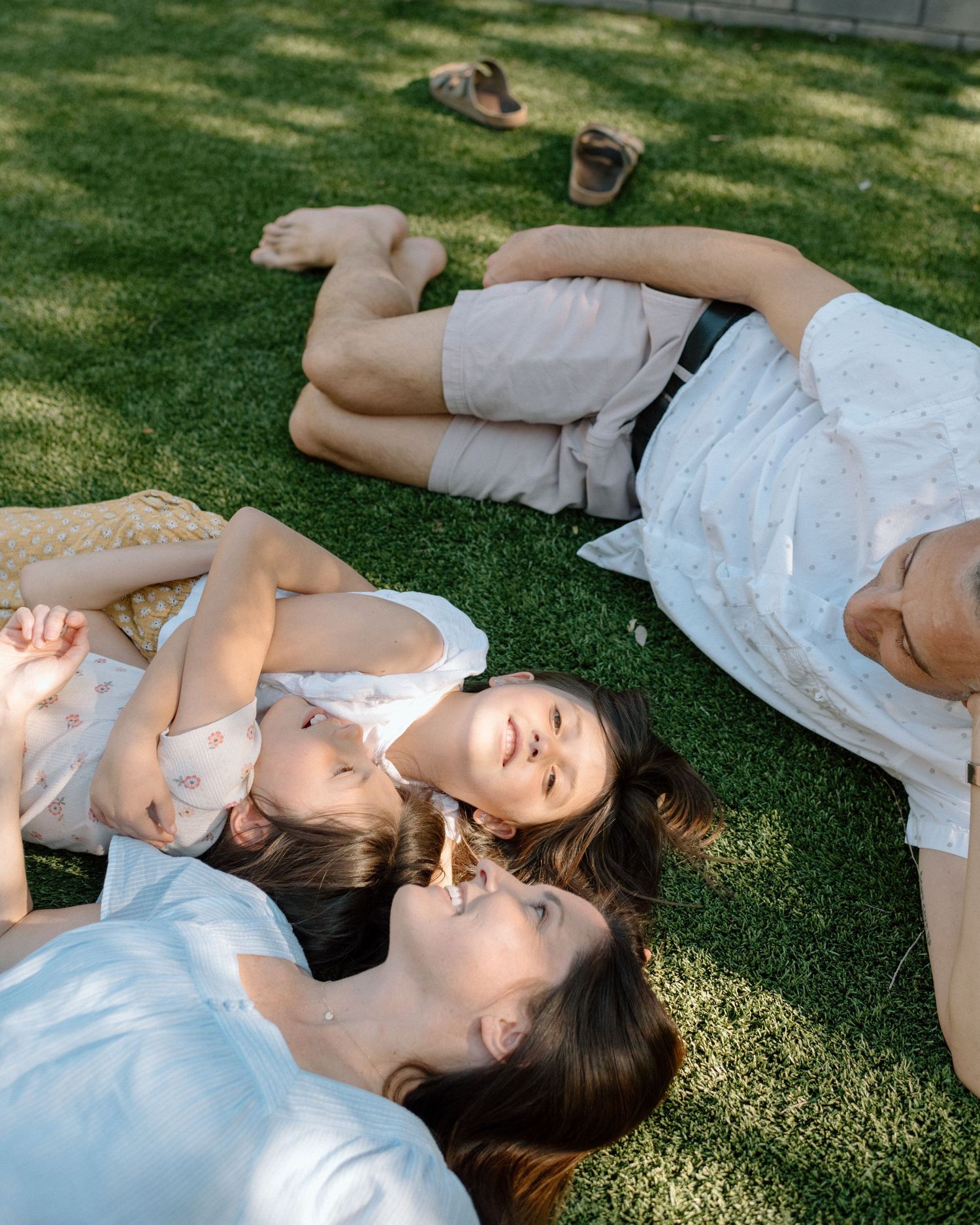 Mother and two young daughters lying together on green grass in warm sunlight.