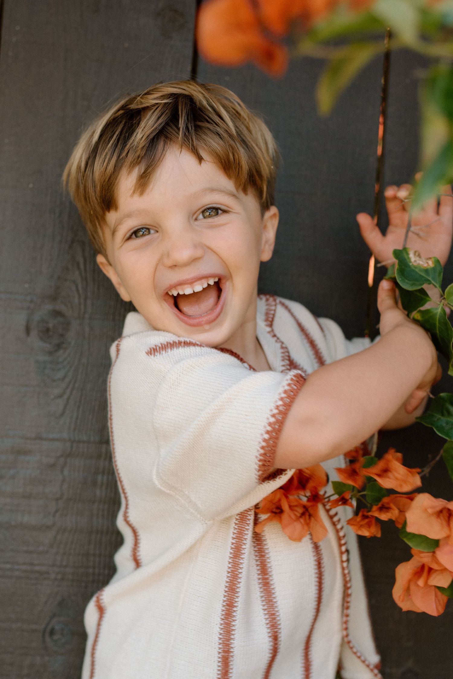 A joyful young boy laughing and reaching for orange flowers against a rustic wooden backdrop.