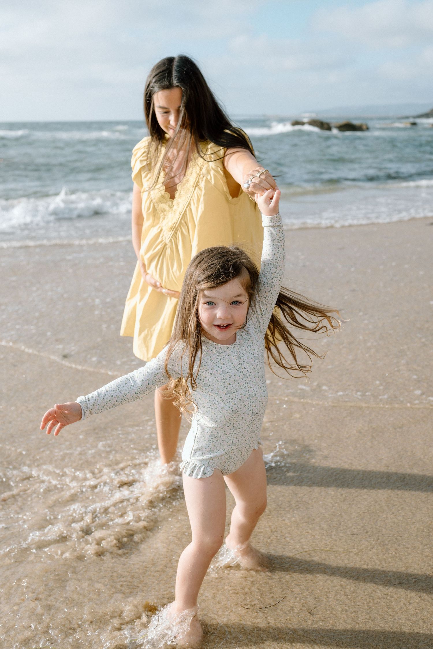 A young girl splashes in ocean waves while an older girl in a yellow dress holds her hand on a sunny beach.