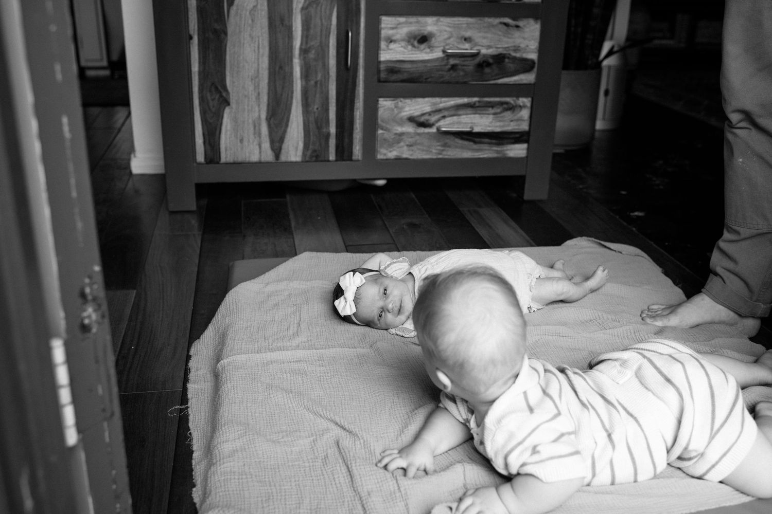 Black and white photo of two babies lying on a blanket on a hardwood floor near wooden furniture.