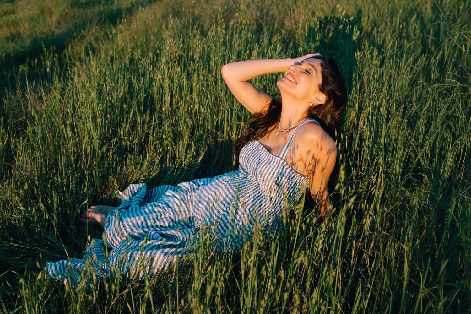 Woman in striped dress relaxes in tall green grass during golden hour sunlight.