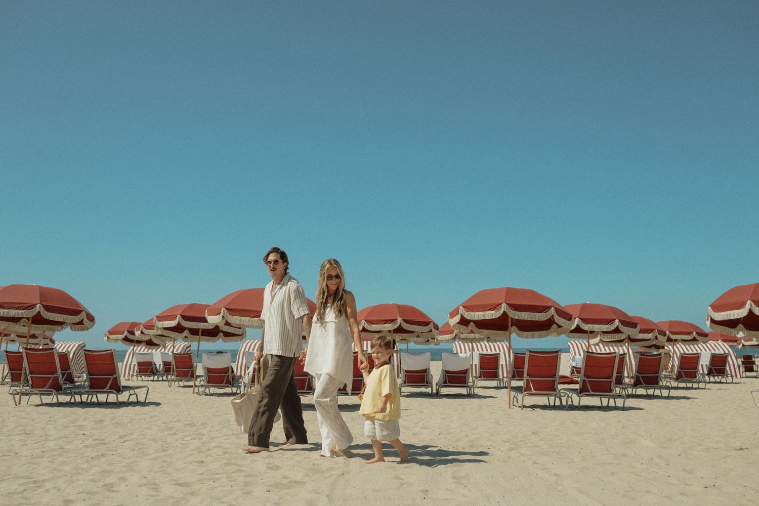 Family of three standing on a sandy beach surrounded by red striped umbrellas and lounge chairs under a clear blue sky.