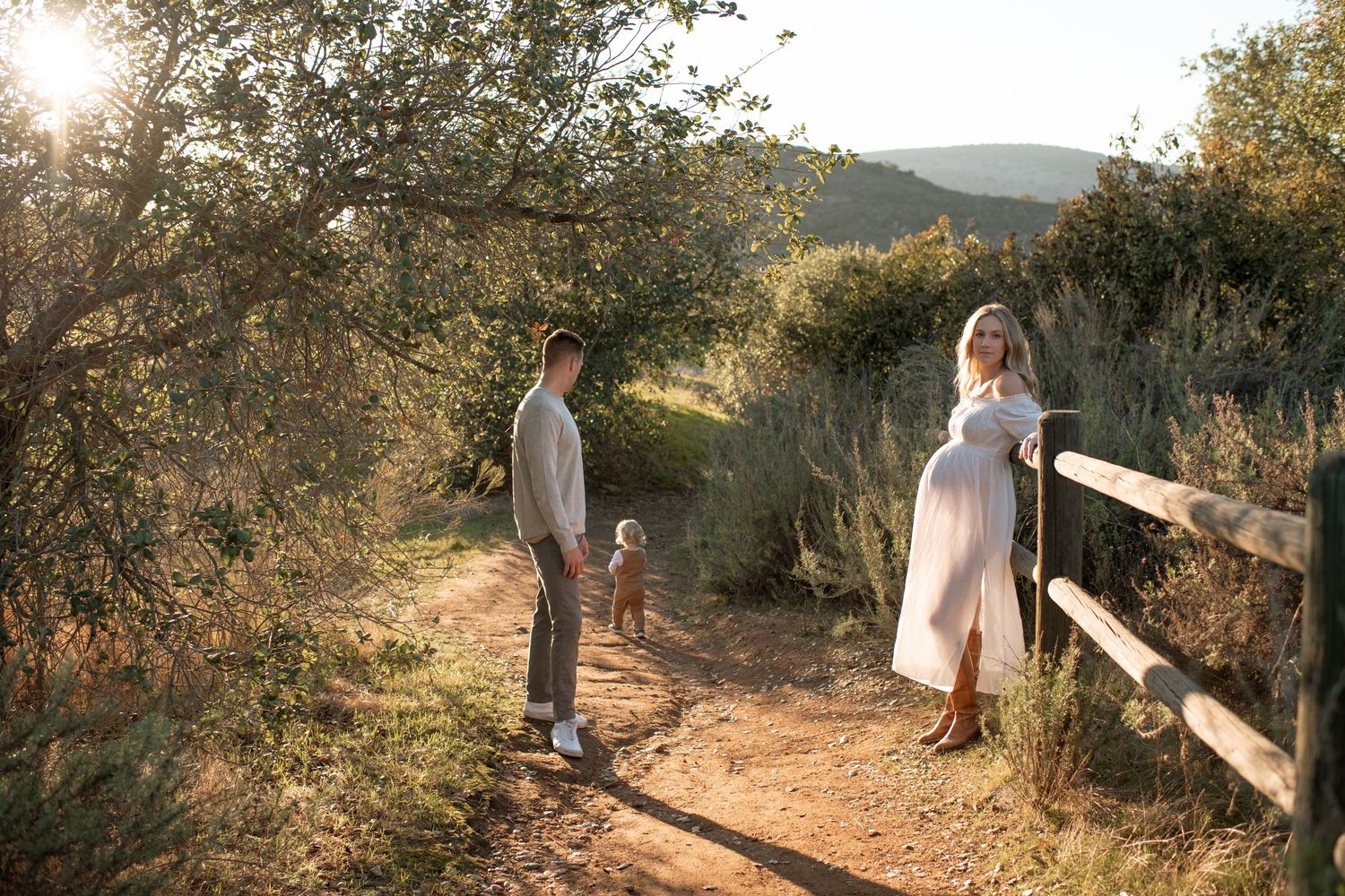 Family walking on a sunlit dirt path surrounded by trees and hills during maternity shoot.