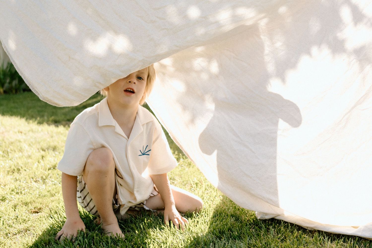 Young boy sitting on grass under a billowing white sheet in sunny backyard.