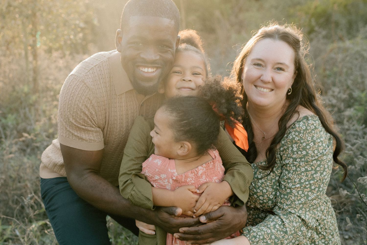 Smiling interracial family of four posing together outdoors in golden sunlight with two young daughters.