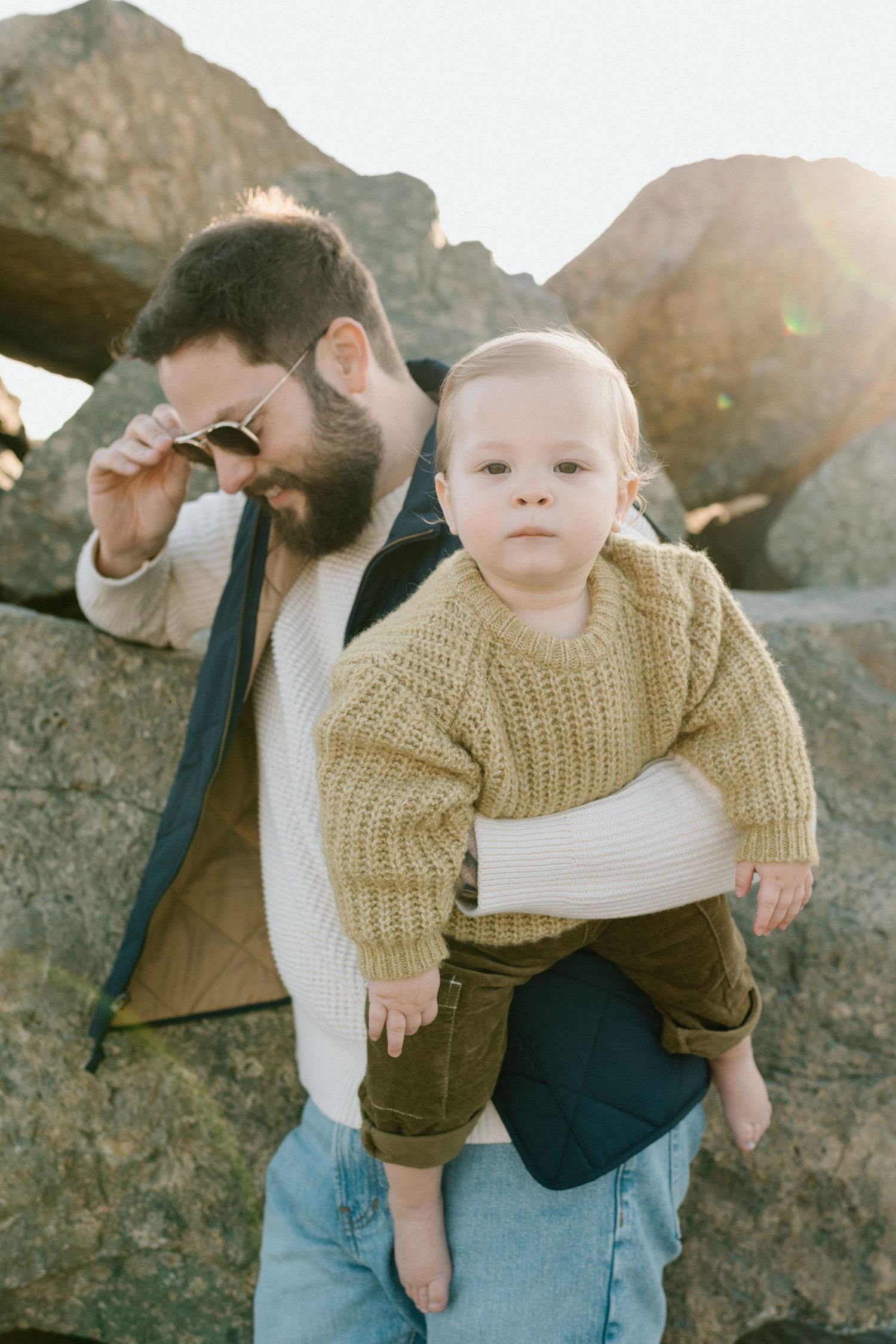 A bearded father holds his baby in a knit sweater near large rocks in golden backlight.