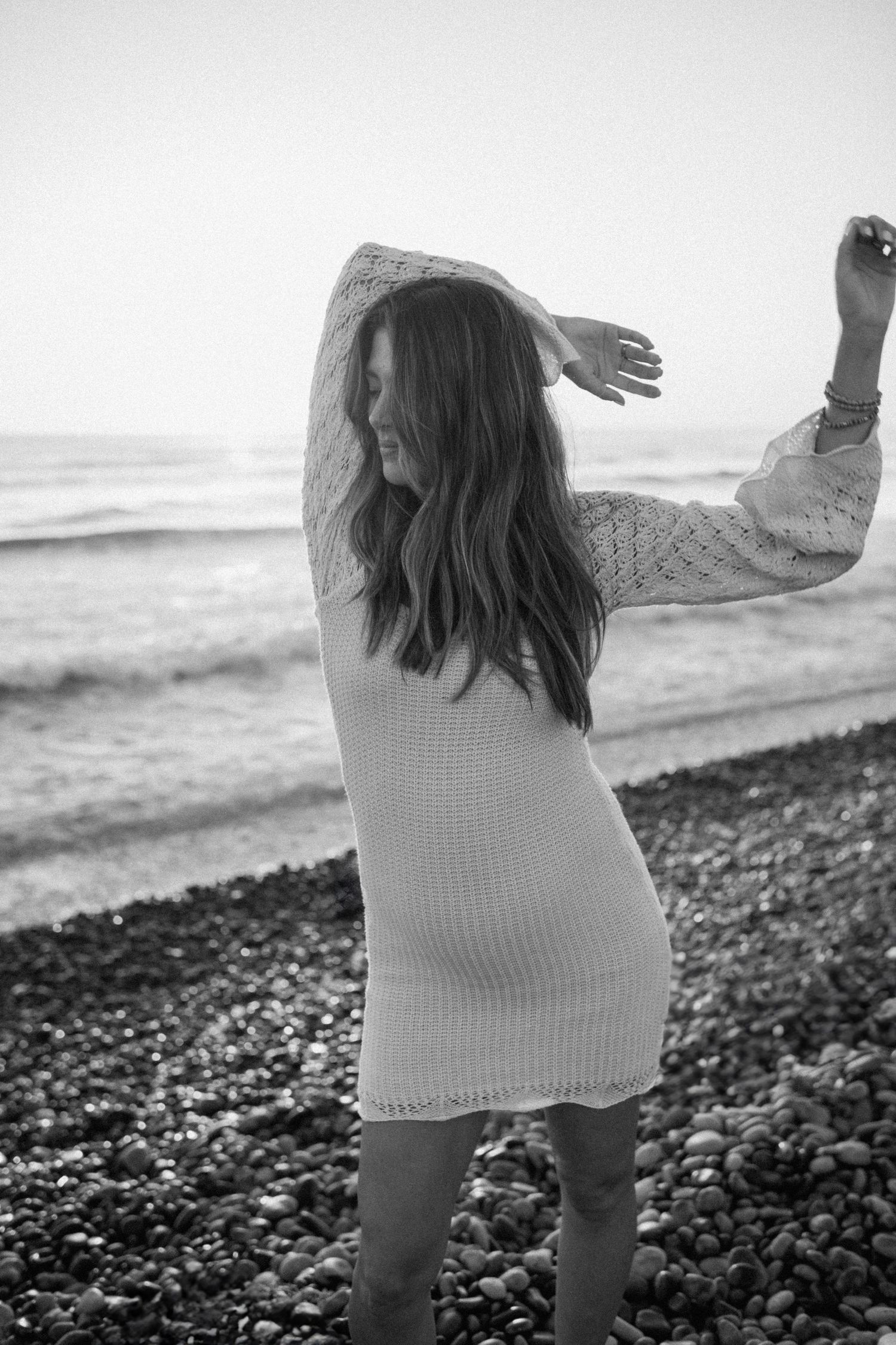 Black and white photo of person wearing a sweater dress dancing on a rocky beach at the ocean.
