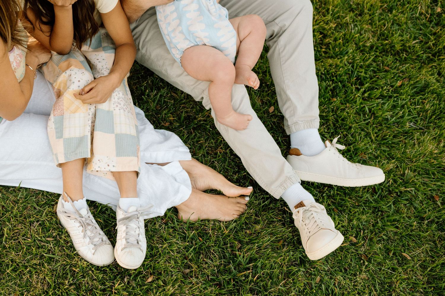 Family sitting on green grass, showing legs and feet in white sneakers, pastel outfits, with a baby's bare feet visible.