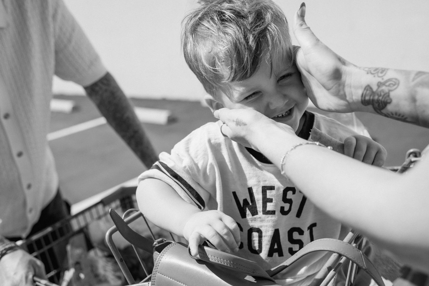 A young boy in a West Coast shirt laughs while being held in a shopping cart by tattooed adults outdoors.