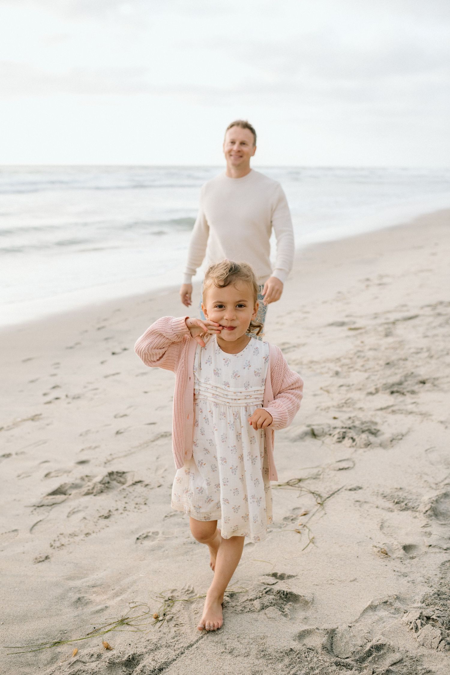 A young girl in a pink cardigan and floral dress walks barefoot on a sandy beach with her father behind her.