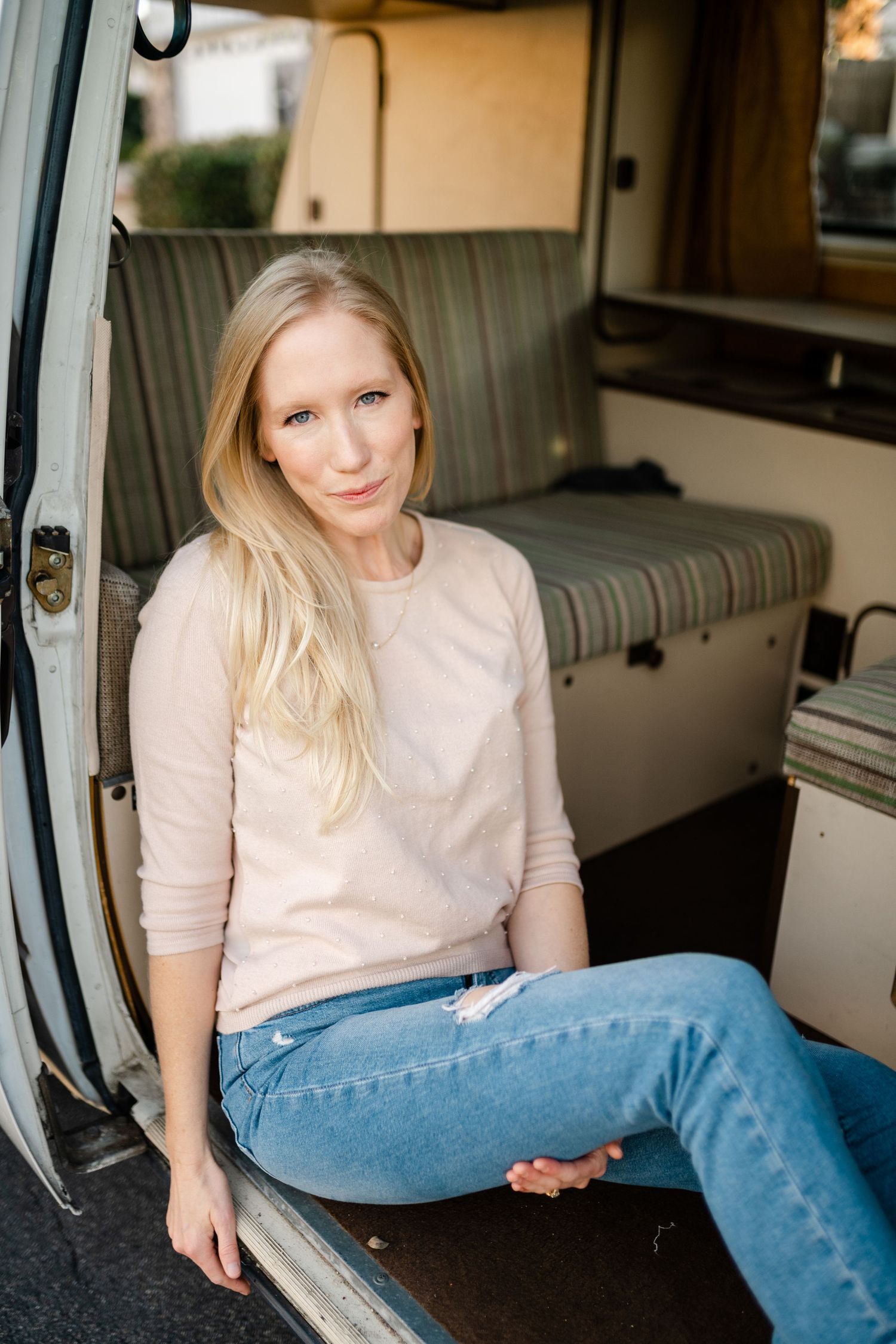 Blonde woman in pink sweater and jeans sitting in open camper van doorway smiling at camera.