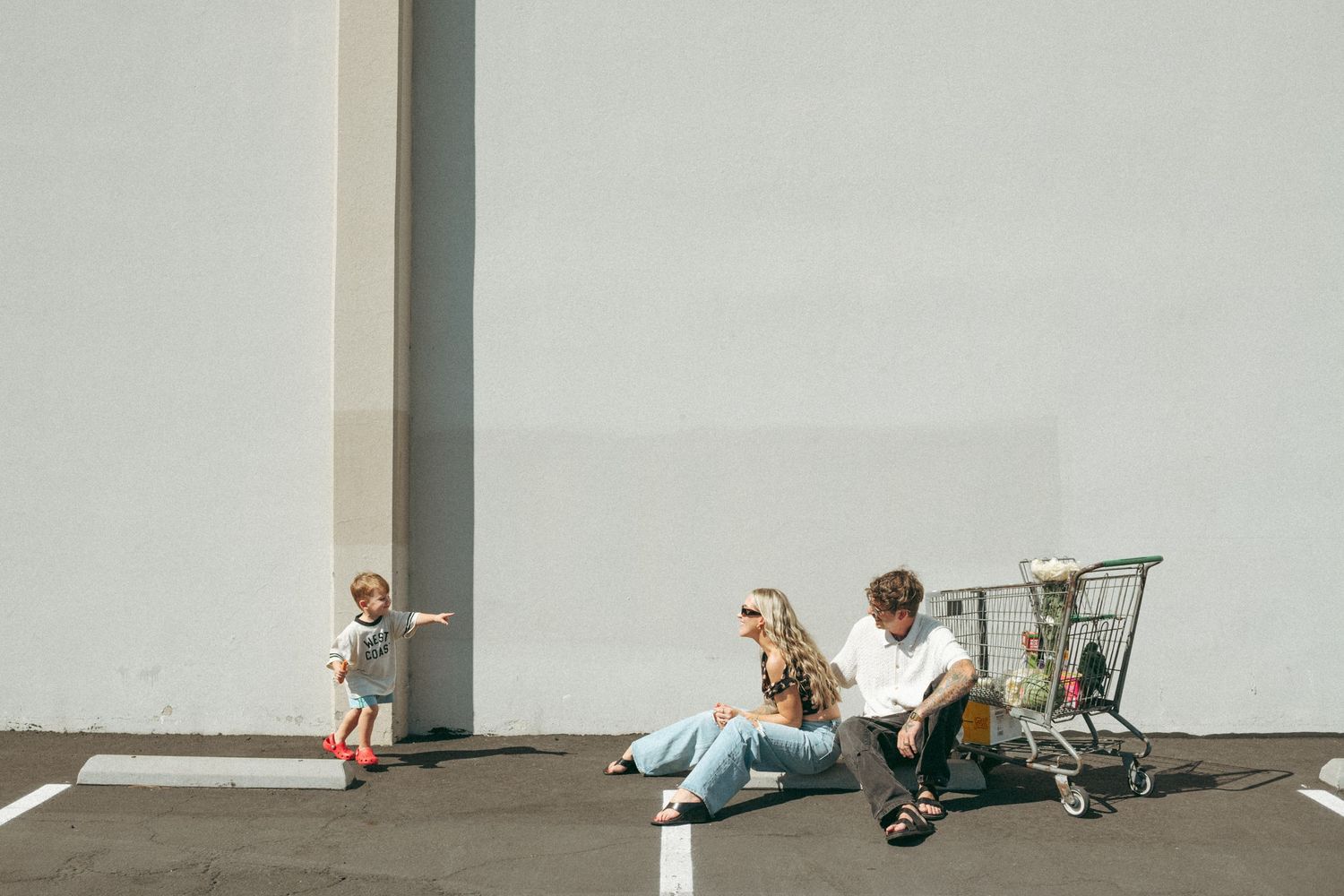 Family relaxing in a sunny parking lot beside a shopping cart while a young child plays nearby against a white wall.